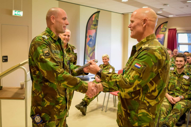 Zwei Männer in Militäruniformen geben sich die Hand vor einer sitzenden Gruppe, mit einem Tisch, Geländer, Fahnen und diversen Gegenständen im Hintergrund, was auf eine deutsche Armee-Versammlung hinweist.