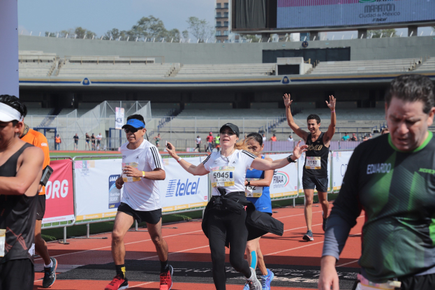Gruppe von Läufern auf einer Tartanbahn in einem Stadion mit Bannern, einem Metallzaun, einem Display, einem Gebäude, Bäumen und einer bewölkten Himmel; einige Läufer tragen Sneaker und Hüte.