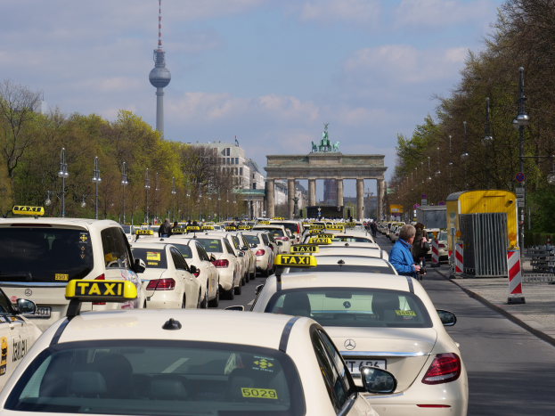 Eine belebte Straße in Berlin, Deutschland, mit vielen parkenden Taxis, Fußgängern auf dem Gehweg, Laternenpfählen, Bäumen und Gebäuden im Hintergrund, mit einem fernen Bogen mit Statuen und einem Turm unter einem bewölkten Himmel.