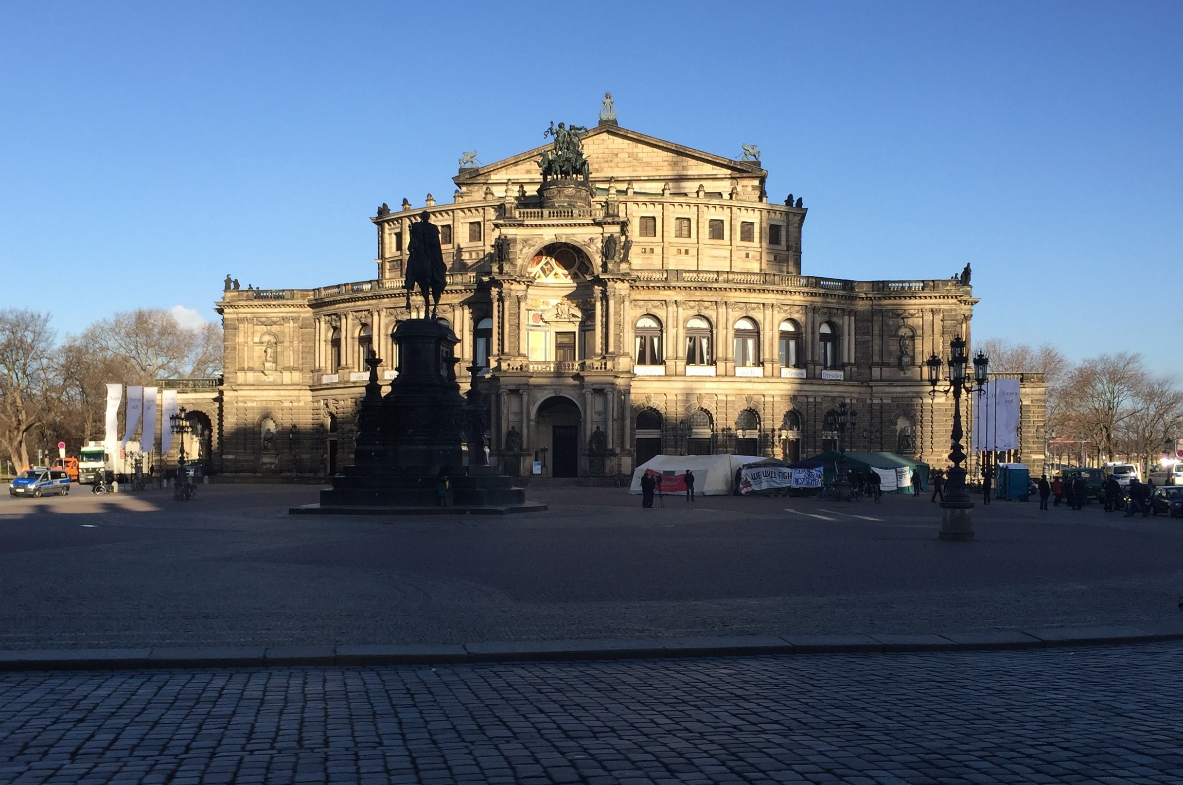 Großes beige Berliner Dom-Gebäude mit einem prominenten Kuppel, eine Statue davor, umgeben von Straßenlaternen, Lichtern, Fahrzeugen, Menschen, Zelten, Bäumen und einem bewölkten Himmel.
