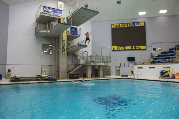 Großer Hallenbad mit schwimmenden Menschen, Treppe mit Geländer, Banner mit Text, Person beim Sprung ins Wasser, Stühle rechts, Bildschirm an der Wand und Deckenbeleuchtung.