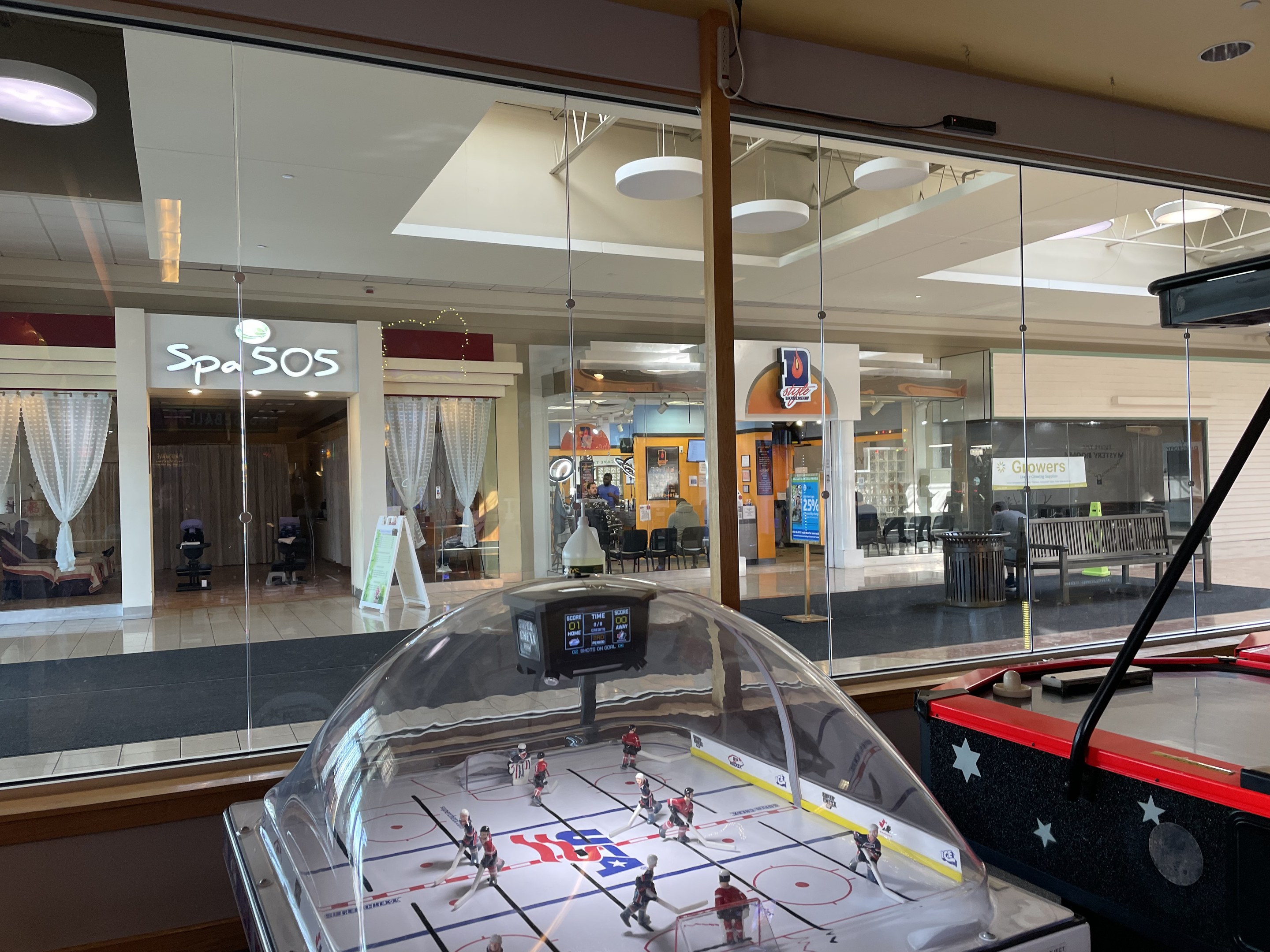 Eine Eishalle in einem Einkaufszentrum, umgeben von Glaswänden mit Menschen, Möbeln und anderen Gegenständen, die von außen sichtbar sind, und einem Air-Hockey-Tisch auf der rechten Seite.
