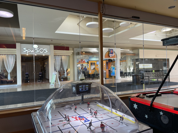 Eine Eishalle in einem Einkaufszentrum, umgeben von Glaswänden mit Menschen, Möbeln und anderen Gegenständen, die von außen sichtbar sind, und einem Air-Hockey-Tisch auf der rechten Seite.