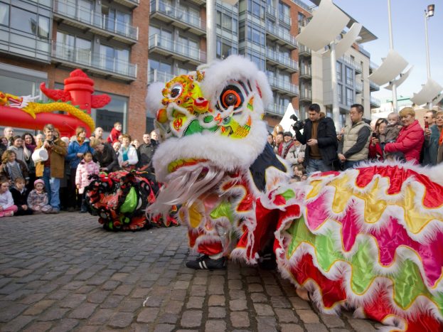 Ein farbenfrohes chinesisches Neujahrsfest in Amsterdam mit einem Löwen Tanz im Vordergrund und einer Zuschauermenge, einige halten Kameras, vor einem Hintergrund aus Gebäuden, Laternenmasten und einem klaren blauen Himmel.