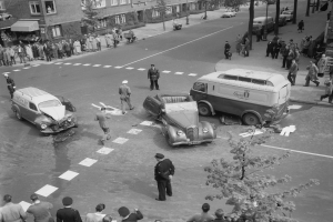 Eine Schwarz-Weiß-Szene eines Autounfalls an einer Straßenkreuzung, mit mehreren Fahrzeugen, darunter ein Transporter, einer Gruppe von Menschen, die sich darum versammeln, und Gebäuden mit Fenstern, Bäumen, Pfosten und Texttafeln im Hintergrund.