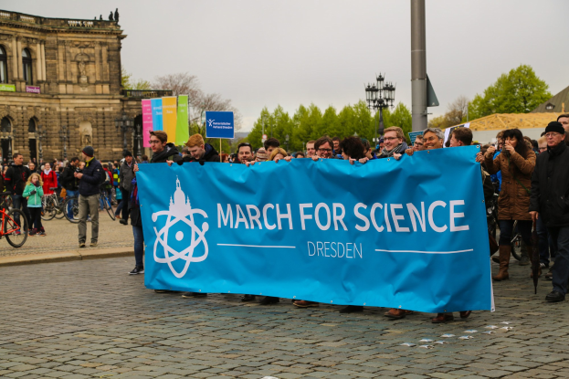 Eine Gruppe von Menschen hält eine "March for Science Dresden"-Plakette vor einem Gebäude mit Fahrrädern, Bäumen und Laternenmasten, unter einem sichtbaren Himmel.