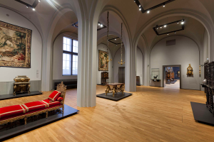 Ein geräumiger Raum im Kunsthistorischen Museum in Wien, Österreich, mit einem Sofa mit Kissen, Tischen mit verschiedenen Gegenständen, Säulen, Fenstern und Deckenleuchten sowie zahlreichen paintings an den Wänden.