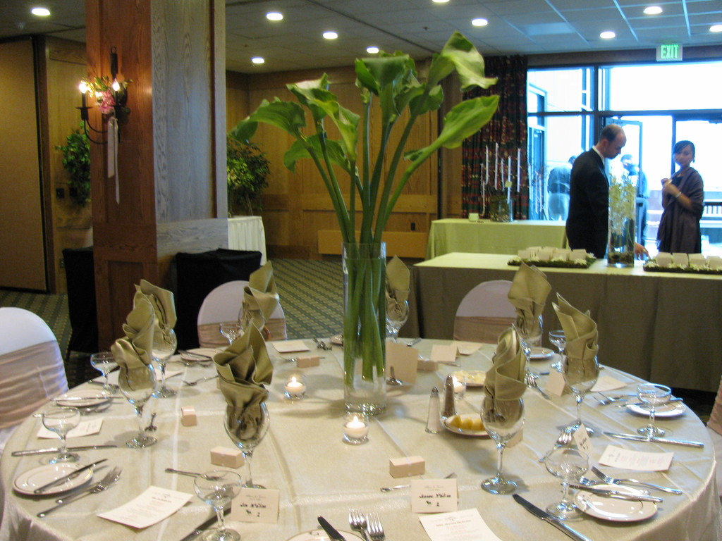 Ein Tisch mit einer weißen Decke, Tellern, Besteck und einem Glas, umgeben von Stühlen, mit einem grünen Tisch dahinter; ein Mann in einer schwarzen Jacke und eine Frau in der Nähe einer Tür mit zwei Männern dahinter.