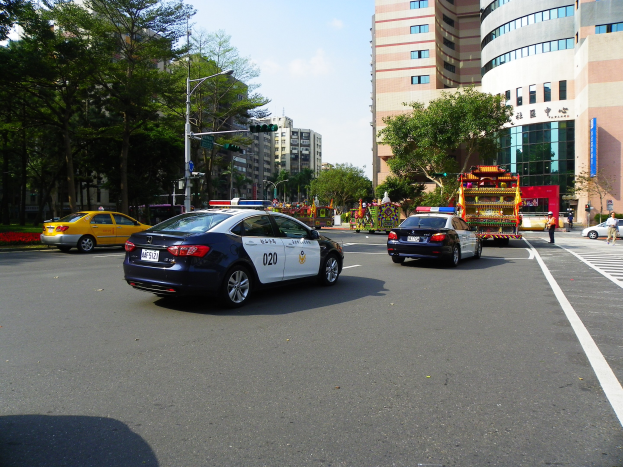 Polizeiauto fährt auf einer Straße mit hohen Gebäuden, Bäumen, Laternen, Verkehrsschildern und Menschen unter einem bewölkten Himmel.