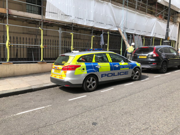 Ein Polizeiwagen steht an der Seite einer Straße neben einem Gebäude, daneben stehen zwei Helme tragende Personen; im Vordergrund sind ein Geländer und ein Pfahl zu sehen, im Hintergrund ein weißes Tuch über das Gebäude drapiert, was auf eine polizeiliche Untersuchung nach einem Messerangriff in London hindeutet.