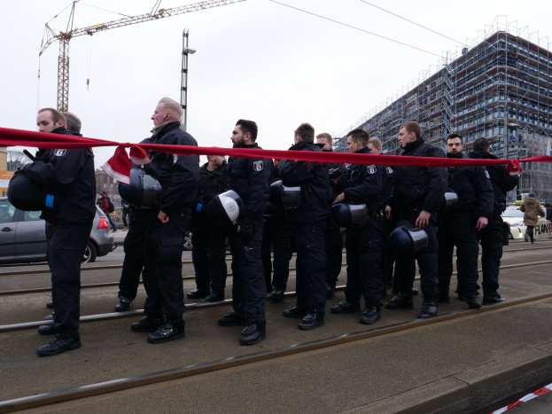 Eine Gruppe von Polizisten in schwarzen Uniformen und Helmen steht in einer Reihe auf einem Bahngleis, mit einem roten Band davor, vor einer Kulisse aus Fahrzeugen, Fußgängern, Gebäuden und einem klaren Himmel.