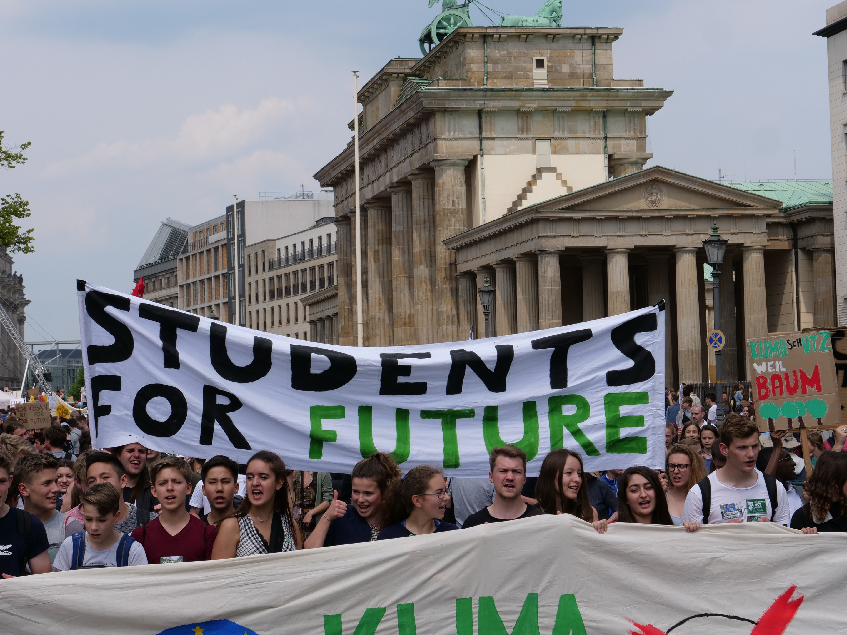 Gruppe von Schülern marschiert in Berlin mit einem bunt bemalten "Students for Future"-Schild an Gebäuden, Bäumen und Himmel vorbei.