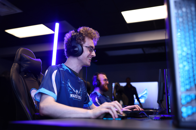 Ein Mann in einem Headset und Brille sitzt an einem Schreibtisch mit einem Computer-Monitor, Tastatur und Maus und spielt ein Spiel in einem Raum mit einer anderen Person und Deckenleuchten.