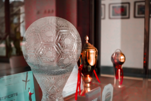 Glastrophäe auf einem Glastisch umgeben von anderen Trophäen und einem Zertifikat, mit gerahmten Gegenständen an der Wand im Hintergrund.