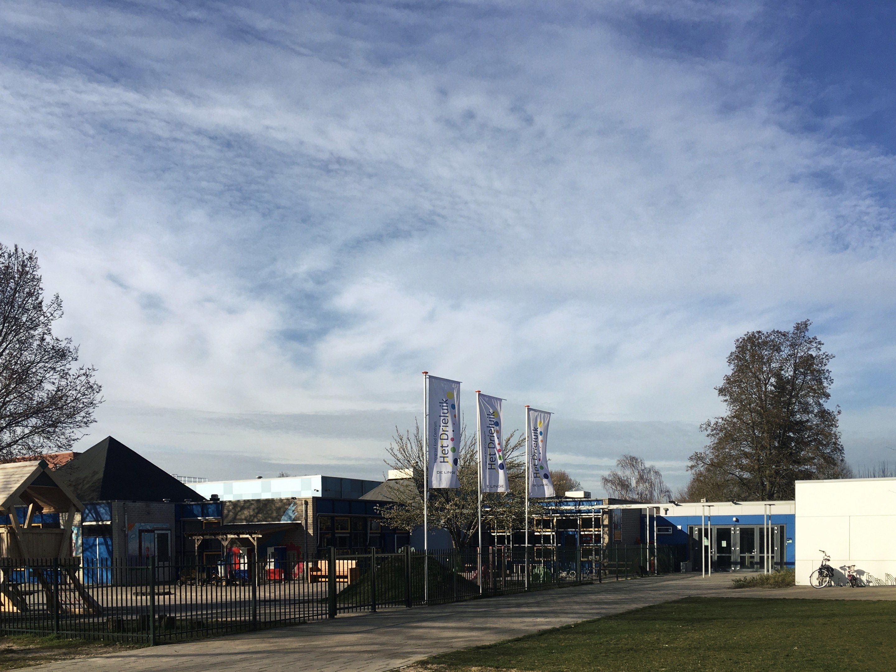 Großes Gebäude mit Metallzaun, Rasenfläche, Weg, Bäumen, Pfählen, Bannern, Fahrrad, nahen Häusern und bewölktem Himmel, identifiziert als neuer Spielbereich für Kinder an der Schule.