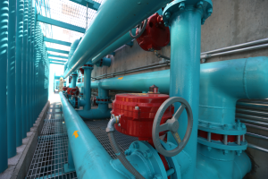 Eine große Gruppe von blauen Rohren und Ventilen in einem Geböude, mit einem roten Ventil in der Mitte, Metallstangen links und einer Wand im Hintergrund, wahrscheinlich zeigt das Bild den Bau einer Gasleitung.