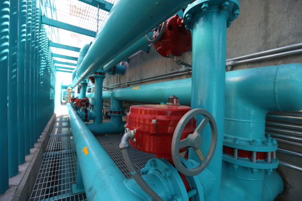 Eine große Gruppe von blauen Rohren und Ventilen in einem Geböude, mit einem roten Ventil in der Mitte, Metallstangen links und einer Wand im Hintergrund, wahrscheinlich zeigt das Bild den Bau einer Gasleitung.