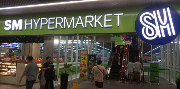 Gruppe von Menschen vor einem hell erleuchteten "SM Hypermarket"-Laden mit Deckenbeleuchtung, Sichttafeln und Topfpflanzen drinnen.