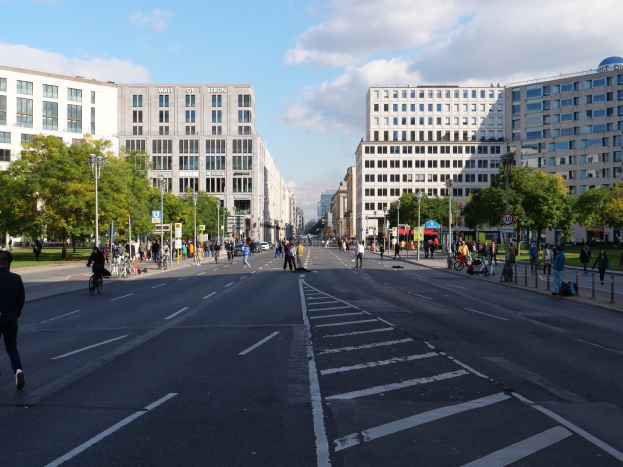 Eine belebte Stadtstraße in Berlin, Deutschland, mit Fußgängern und Radfahrern auf der Straße, umgeben von hohen Gebäuden, Bäumen, Straßenlaternen und Schildern unter einem bewölkten Himmel.