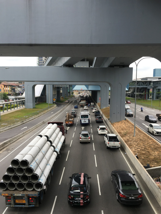 Eine stark befahrene Autobahn mit dichtem Verkehr unter einer Brücke, umgeben von Gras, Bäumen, Gebäuden, Polen und Straßenlaternen unter einem klaren Himmel.