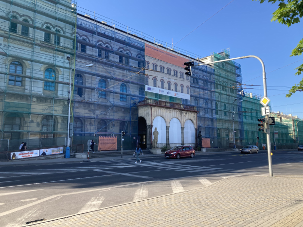 Ein großes Gebäude mit Gerüst, das renoviert wird, umgeben von Fahrzeugen, Passanten, Ampeln, Schildern, Straßeninfrastruktur, Bäumen und einem klaren blauen Himmel.