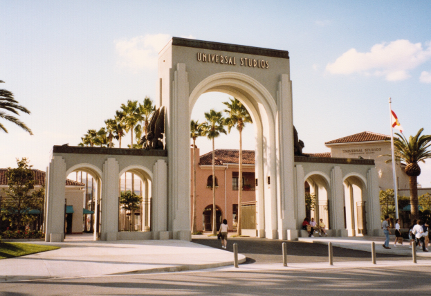 Eingang von Universal Studios Hollywood mit einem Bogen, Statue, Menschen, Fahnenmast, Gebäuden, Bäumen und einem bewölkten Himmel.