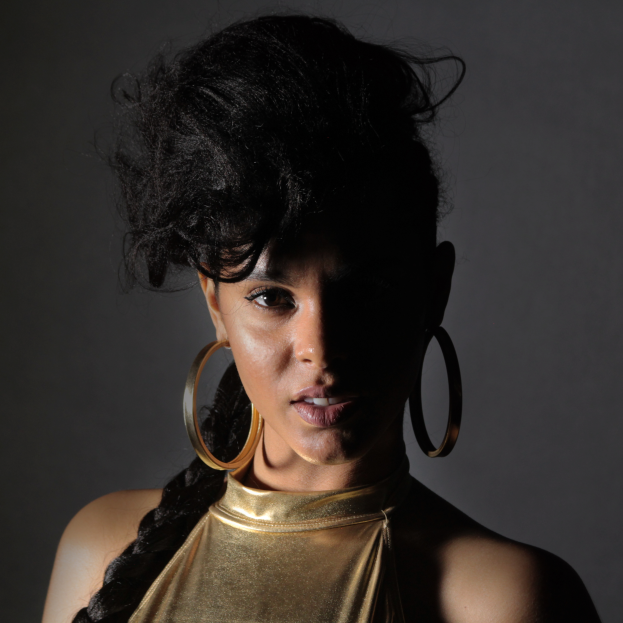 Eine Frau in einem goldenen Kleid und großen Kreolen steht selbstbewusst vor einer Wand, ihr Haar ist zu einem glatten Bob gestylt, sie trägt dezentes Make-up mit pinkem Rouge und glänzenden Lippen, blickt direkt in die Kamera mit einem starken, entschlossenen Ausdruck.