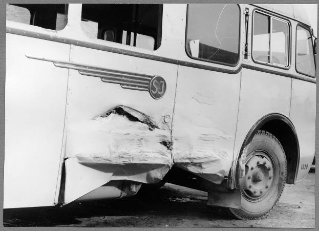 Schwarzes und weißes Foto eines verunglückten Busses mit eingedrückter Front, zersplitterten Fenstern und geöffneter Fahrertür, die den Innenraum zeigt.