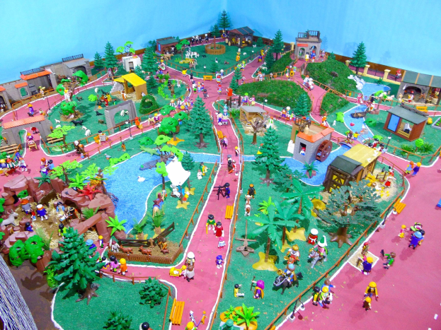 Ein Miniaturmodell eines Zoos mit Besuchern, umgeben von Bäumen, Häusern, Pflanzen, Gras, einem Zaun und einer Hintergrundwand, mit Text unten.