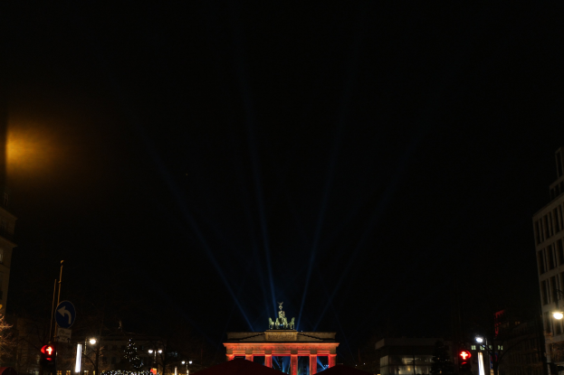 Brandenburger Tor in Berlin bei Nacht, beleuchtet von Straßenlaternen, mit umliegenden Gebäuden, Bäumen, Pfählen, Verkehrszeichen, Schildern und Zelten gegen einen dunklen Himmel.