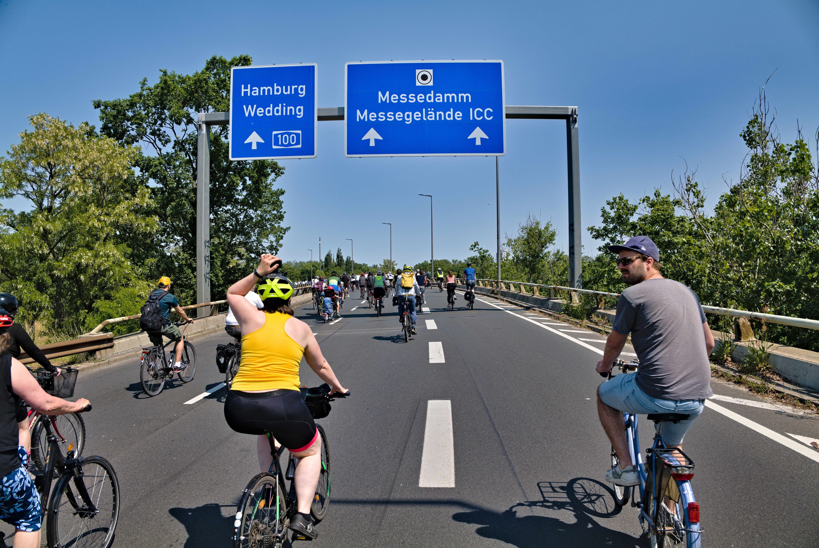 Eine Gruppe von Radfahrern in Helmen fährt eine Straße entlang, die von Bäumen und einem Geländer gesäumt ist, mit Laternenmasten und einem klaren blauen Himmel im Hintergrund; ein Schild oben zeigt eine Radtour in Hamburg an.