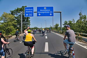 Eine Gruppe von Radfahrern in Helmen fährt eine Straße entlang, die von Bäumen und einem Geländer gesäumt ist, mit Laternenmasten und einem klaren blauen Himmel im Hintergrund; ein Schild oben zeigt eine Radtour in Hamburg an.