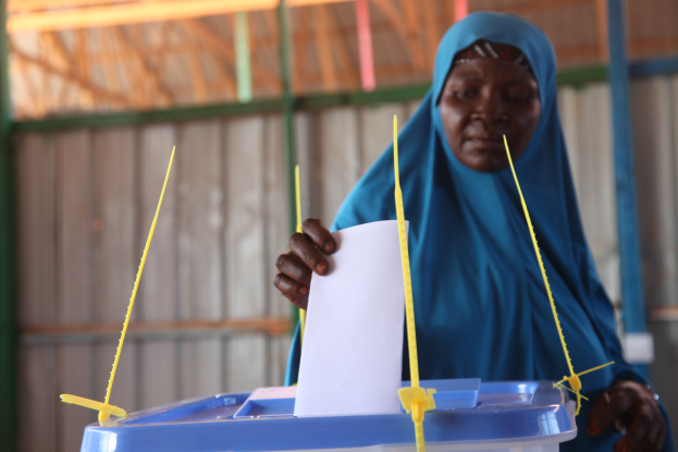 Frau mit blauem Kopftuch, die ein Papier hält und vor einer Kiste mit herausragenden Stöcken steht, vor einer Wand und einem Schuppen im Hintergrund an einer Wahllokal.