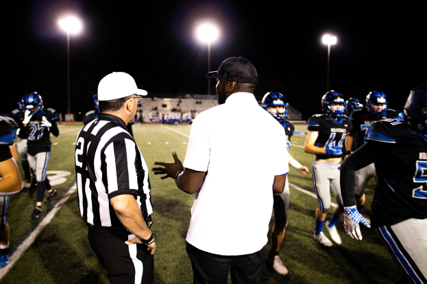 Ein Fußballtrainer in einer Mütze spricht zu seinem Team von Helden auf einem Feld bei Nacht, mit Leuchtmasten und einem dunklen Himmel im Hintergrund.