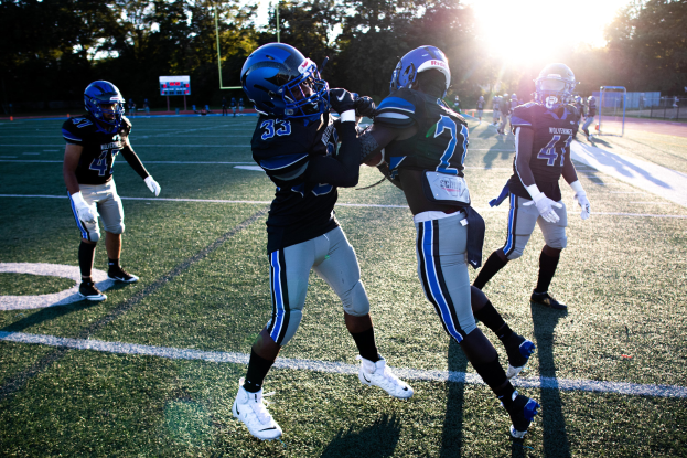 Gruppe junger Männer in Helmen und Schuhen, die Fußball auf einem von Bäumen umgebenen Feld unter einem klaren blauen Himmel spielen.