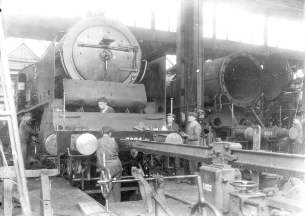 Schwarzes und weißes Foto von Männern, die an einem Zug in einer Fabrik arbeiten, einige halten Werkzeuge, mit einer Leiter auf der linken Seite und einem Schuppen im Hintergrund.