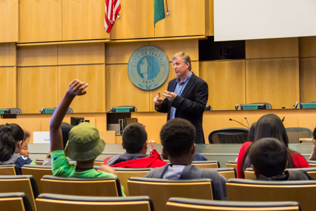 Ein Mann in einem schwarzen Blazer, blauer Hemd und schwarzen Hosen steht vor einer Klasse aufmerksamer Kinder, h├Ąlt ein Mikrofon in der Hand und hat eine h├Âlzerne Wand mit einem Logo und zwei Flaggen hinter sich und einen weißen Bildschirm rechts.