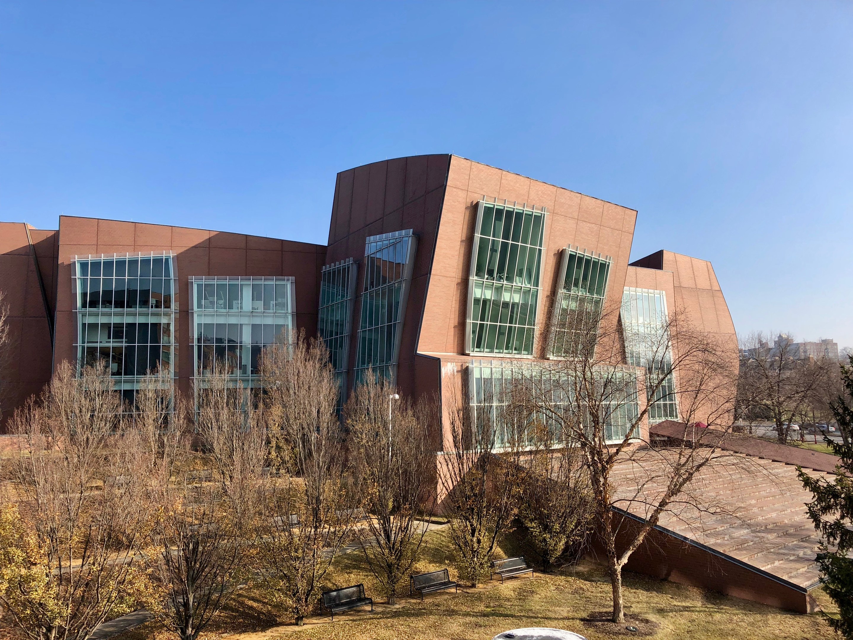 Großes Gebäude mit vielen Fenstern, umgeben von Bäumen, Bänken, Gras und einem Laternenmast, mit anderen Gebäuden und einem klaren blauen Himmel im Hintergrund; Teil des neuen Campus der University of Illinois at Chicago.