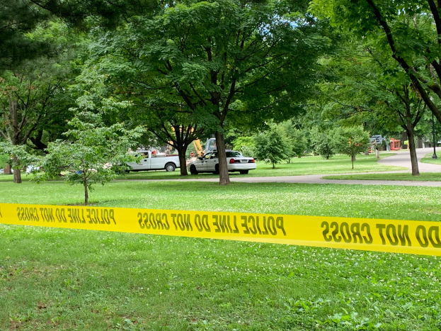 Eine Grasfläche mit gelbem Polizeiabsperrband in der Mitte, umgeben von Bäumen und Fahrzeugen auf der Straße, wahrscheinlich zeigt die Nachwirkung einer Schießerei in einem Park.