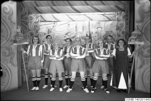 Eine Gruppe strahlender Frauen in einem 1940er Fußballteam posiert auf einer Bühne, jede hält einen Stock in der Hand, mit einer bemalten Wand im Hintergrund.
