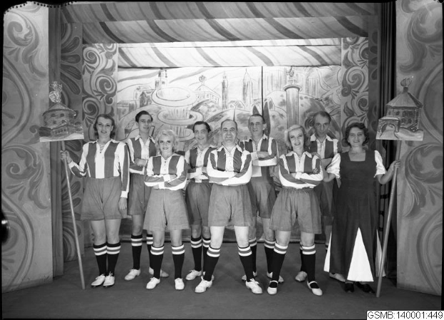 Eine Gruppe strahlender Frauen in einem 1940er Fußballteam posiert auf einer Bühne, jede hält einen Stock in der Hand, mit einer bemalten Wand im Hintergrund.