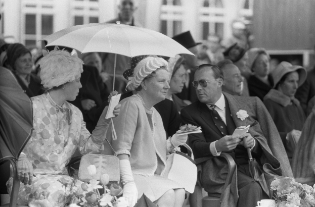 Schwarzes und weißes Foto von Queen Elizabeth II. und Prinz Philip, die mit Regenschirmen auf Stühlen sitzen, umgeben von einer Menge mit Hüten und Büchern, mit einem Gebäude und Blumenarrangements im Hintergrund.