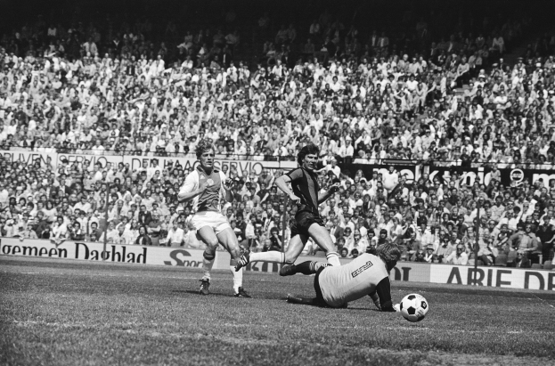 Schwarze-Weiß-Foto von Fußballspielern im Einsatz auf einem Feld mit Zuschauern im Hintergrund und einer "Bundesliga 1978-79 Ajax 1-1 Hannover 96"-Fahne.