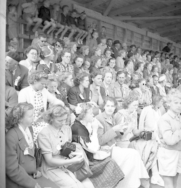 Schwarzes und weißes Foto einer Menge, die in einem Stadion mit einer hölzernen Wand und einem Dach im Hintergrund sitzt, wobei einige Kameras halten.