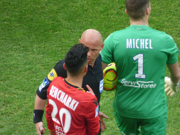 Zwei Fußballspieler und ein Schiedsrichter auf einem grasbewachsenen Feld, wobei einer der Spieler einen Ball hält.