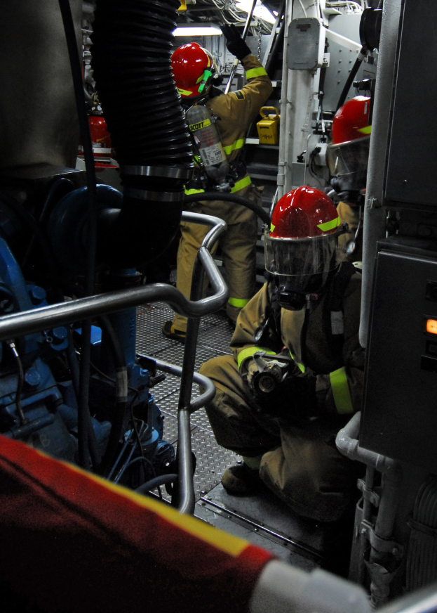 Feuerwehrleute in Helmen und Schutzausrüstung arbeiten an einem Löschfahrzeug mit Rohren und einer Leuchte im Hintergrund.