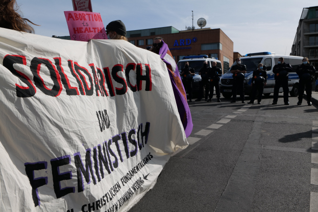 Gruppe von Menschen, die eine Straße entlangmarschieren und ein "Solidarität und Feminismus"-Schild halten, mit parkenden Autos, Gebäuden, einer Schüsselantenne und einem klaren blauen Himmel im Hintergrund.
