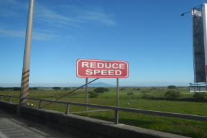 Eine Straße mit einem roten "Reduzieren Sie die Geschwindigkeit"-Schild, ein Pfahl mit einer Tafel und ein Geländer; Bäume, Gras, Berge und Wolken im Hintergrund.