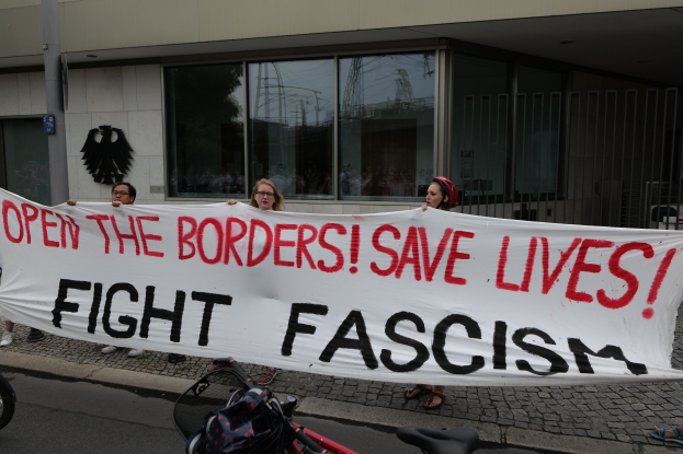 Eine Gruppe von Menschen mit einem Transparent mit der Aufschrift "Öffnet die Grenzen, rettet Leben, bekämpft Faschismus" vor einem Gebäude mit Glasfenstern und einem Mast, mit einem Fahrrad und einer Tasche auf der Straße.