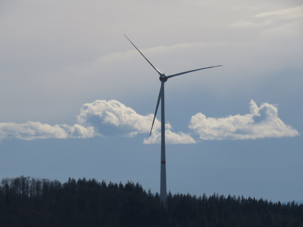 Windkraftanlage in bewaldeter Gegend mit bewölktem Himmel und Bergen im Hintergrund.